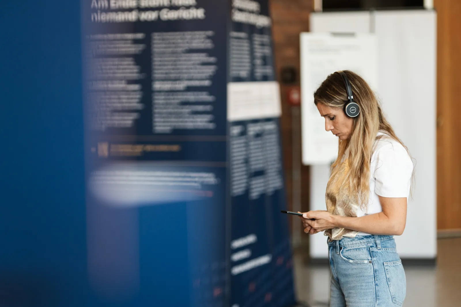 Besucherin mit Kopfh&ouml;rern und Smartphone vor einer Ausstellungstafel